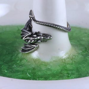 May include: A silver dragon ring with a coiled tail and wings. The ring is displayed on a white ring holder with green glitter.