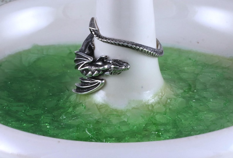 May include: A silver dragon ring with a coiled tail and wings. The ring is displayed on a white ring holder with green glitter.