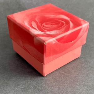 May include: A small, square gift box with a red background and a white rose design. The box is closed and has a pink bottom.