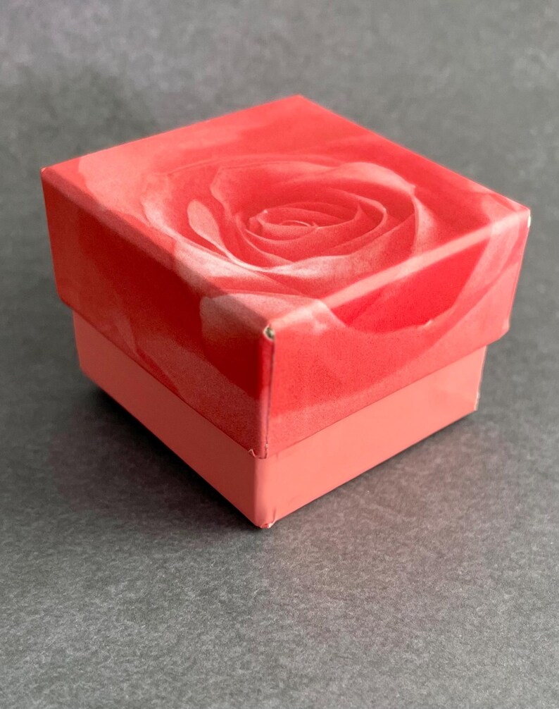 May include: A small, square gift box with a red background and a white rose design. The box is closed and has a pink bottom.