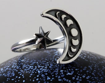 May include: A silver ring with a crescent moon design featuring phases of the moon and a star. The ring is on a black background with blue speckles.