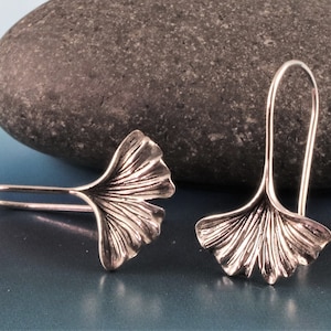 May include: A pair of silver-colored earrings, each shaped like a ginkgo leaf with detailed veining. The earrings are displayed against a blue and gray gradient background, with a dark gray stone in the background.