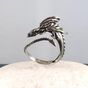 May include: A silver dragon ring with a detailed design. The dragon is curled around the ring band, with its wings spread out.