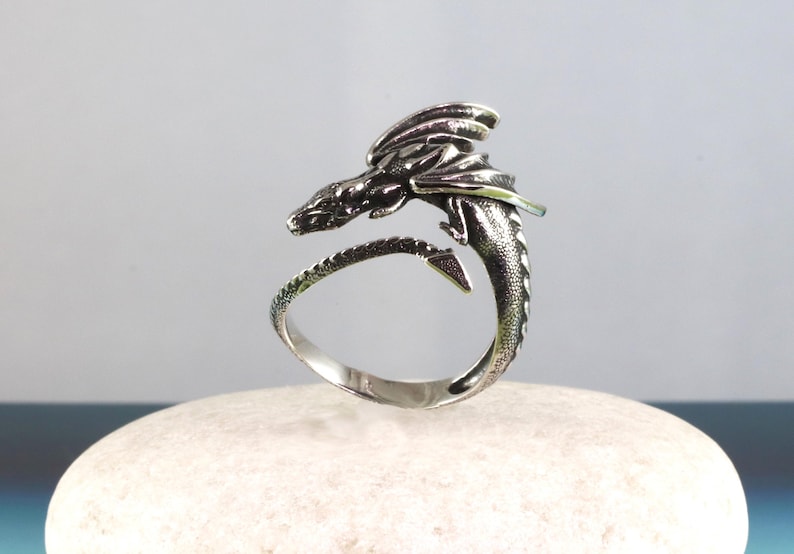 May include: A silver dragon ring with a detailed design. The dragon is curled around the ring band, with its wings spread out.