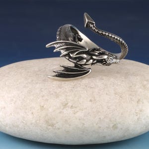 May include: A silver dragon ring with a curled tail and open wings. The ring is sitting on a white stone.