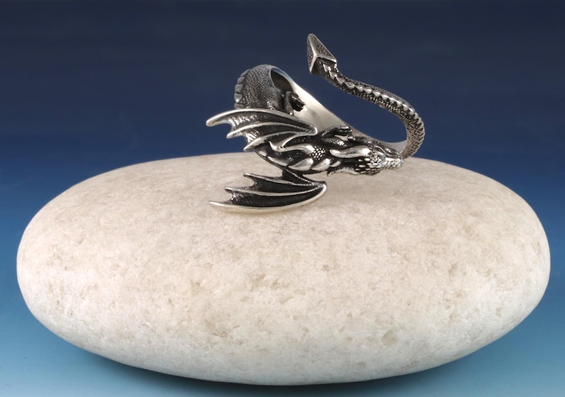 May include: A silver dragon ring with a curled tail and open wings. The ring is sitting on a white stone.