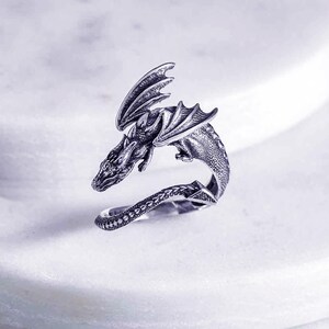 May include: A silver dragon ring with a detailed design. The dragon is curled around the ring band, with its wings spread out.
