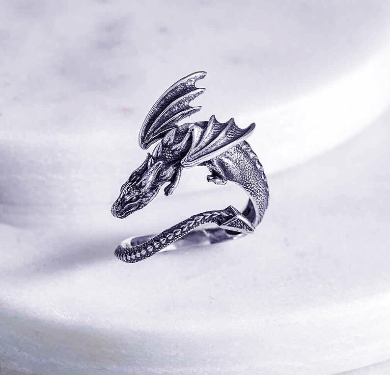 May include: A silver dragon ring with a detailed design. The dragon is curled around the ring band, with its wings spread out.