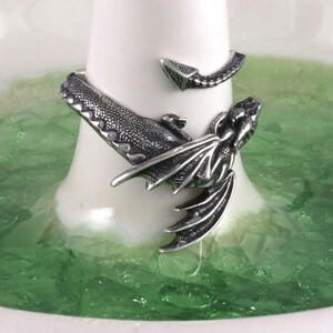 May include: A silver dragon ring with a detailed design. The ring is wrapped around a finger and the dragon's wings are spread out.