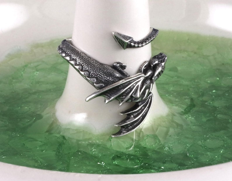 May include: A silver dragon ring with a detailed design. The ring is wrapped around a finger and the dragon's wings are spread out.