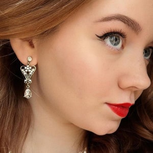 Op de afbeelding: Een oorbel in antieke stijl met een goudkleurig metalen frame en heldere kristallen accenten. De oorbel heeft een hartvormig ontwerp met een druppelvormige kristallen hanger. De vrouw op de foto heeft rode lippenstift en eyeliner.