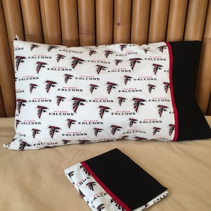 May include: A white pillowcase with a black and red border featuring a repeated pattern of the Atlanta Falcons logo. The pillowcase is folded and placed on a tan surface.