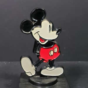 May include: A black and white Mickey Mouse figurine with red shorts stands on a black base with a gold plaque that reads "If you need me, just squeak."