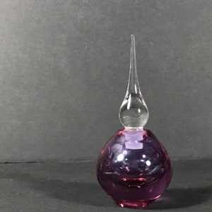 May include: A small, round, purple glass perfume bottle with a clear, elongated, teardrop-shaped glass stopper. The base of the bottle is spherical and translucent, while the stopper features a pointed, tapered top that extends upwards.