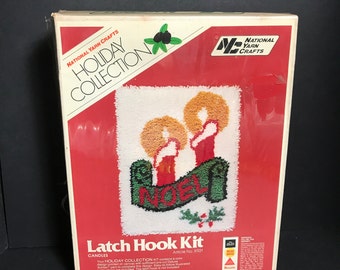 May include: A vintage National Yarn Crafts Holiday Collection latch hook kit. The kit features a design of two candles with orange flames and red bodies, a green banner with the word "NOEL", and holly. The box is red and white with text.