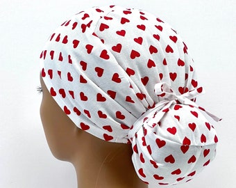 May include: White surgical scrub hat with a red heart pattern. The hat is tied at the back in a bun.