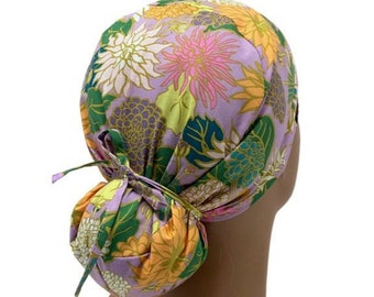 May include: Purple scrub hat with a floral print featuring pink, yellow, and green flowers. The hat has a tie at the back for adjusting the fit.