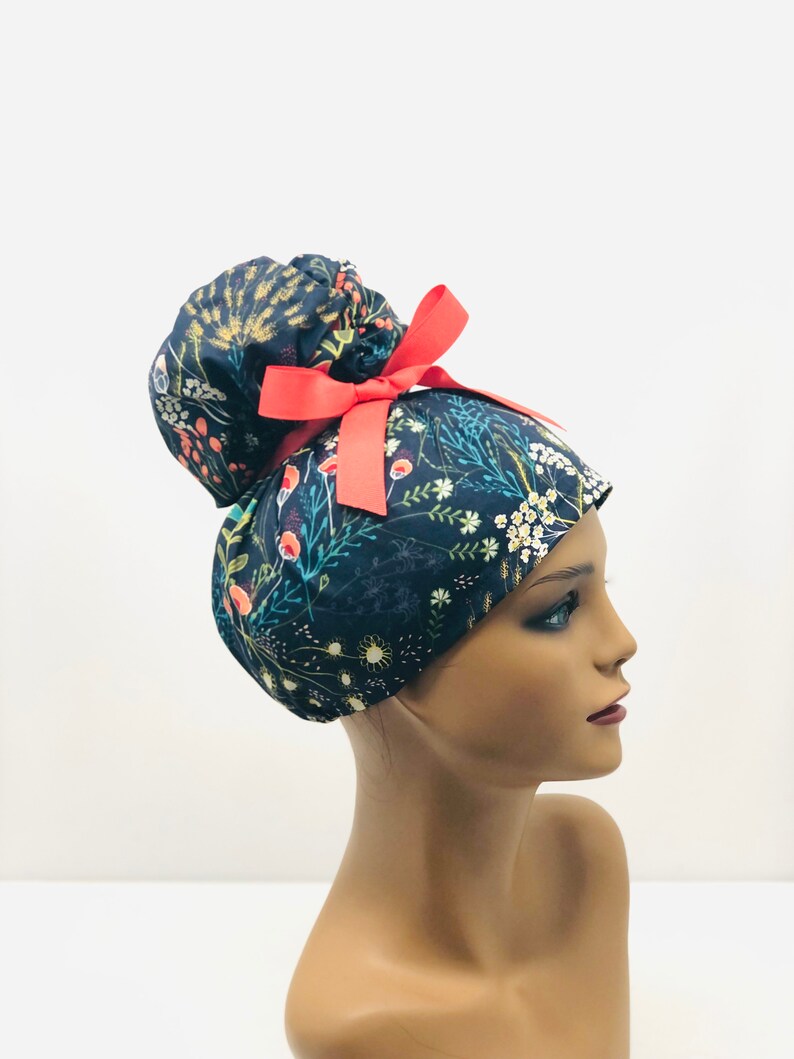 May include: A dark blue scrub hat with a floral print and a red bow. The hat is designed to be worn with a ponytail.