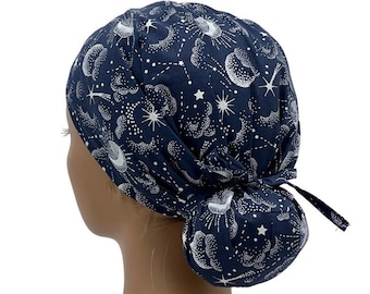 Pode incluir: Um gorro cirúrgico azul marinho com uma estampa branca de estrelas, luas e nuvens. O gorro tem um laço na parte de trás.