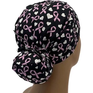 May include: Black scrub hat with a pink ribbon and white heart pattern. The hat has a bun or ponytail holder in the back.