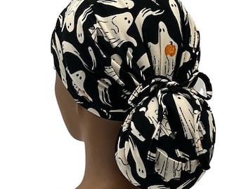 Puede incluir: Un gorro de quirófano negro y blanco con un patrón de fantasmas. El gorro tiene una abertura para una coleta y un lazo en la parte posterior.