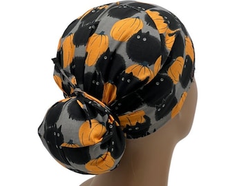 Puede incluir: Un gorro de quirófano gris, negro y naranja con un diseño de gato negro y calabaza naranja. El gorro está atado en un moño en la parte posterior.