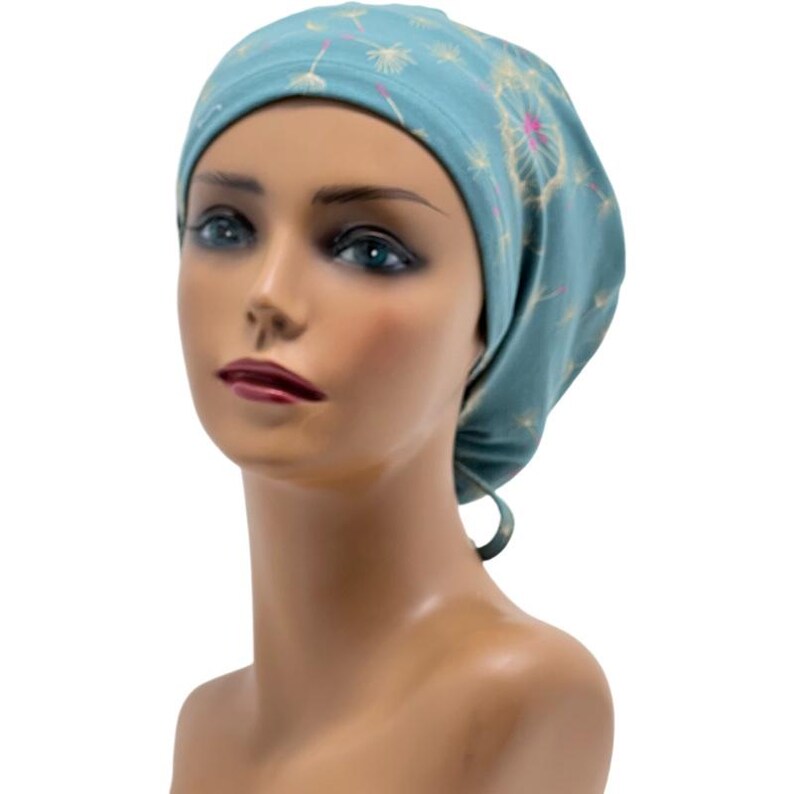 Peut inclure: Un bonnet de chirurgien bleu sarcelle orn&eacute; d'un motif de pissenlits blancs et roses. Le bonnet est nou&eacute; &agrave; l'arri&egrave;re et est pr&eacute;sent&eacute; sur une t&ecirc;te de mannequin. Fabriqu&eacute; en tissu l&eacute;ger et doux.