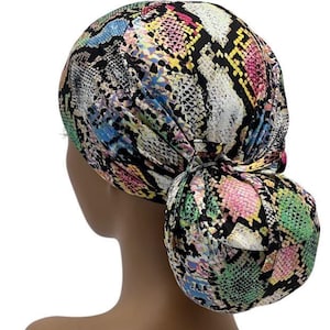 May include: A colorful turban featuring a snakeskin pattern in shades of pink, green, yellow, blue, and black. The turban is tied at the back, with a bun-like knot. The head covering is designed to be worn on the head.