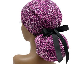 May include: A scrub cap featuring a vibrant purple and black leopard print pattern. The cap includes a gathered back section designed to accommodate hair, secured with a solid black ribbon tied into a bow at the center of the back of the head.