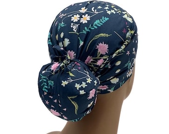 Puede incluir: Un gorro quirúrgico de color azul marino oscuro con un patrón floral repetitivo de margaritas blancas, flores rosas y follaje verde. El gorro incluye una bolsa trasera redondeada para el cabello, asegurada con una cinta de tela en la parte posterior.