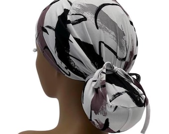 Könnte beinhalten: Eine gemusterte OP-Haube in Weiß, Schwarz und Grau mit einem Pferdeschwanzhalter. Die Haube hat ein Pinselstrich-Design in Grau, Schwarz und Mauve. Sie wird hinten mit einem passenden Stoffband gebunden.