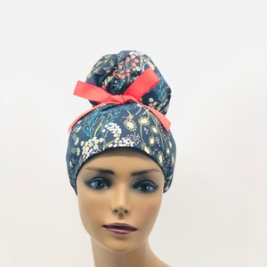 May include: A dark blue scrub hat with a floral print and a bright pink bow. The hat is designed to be worn with a bun.