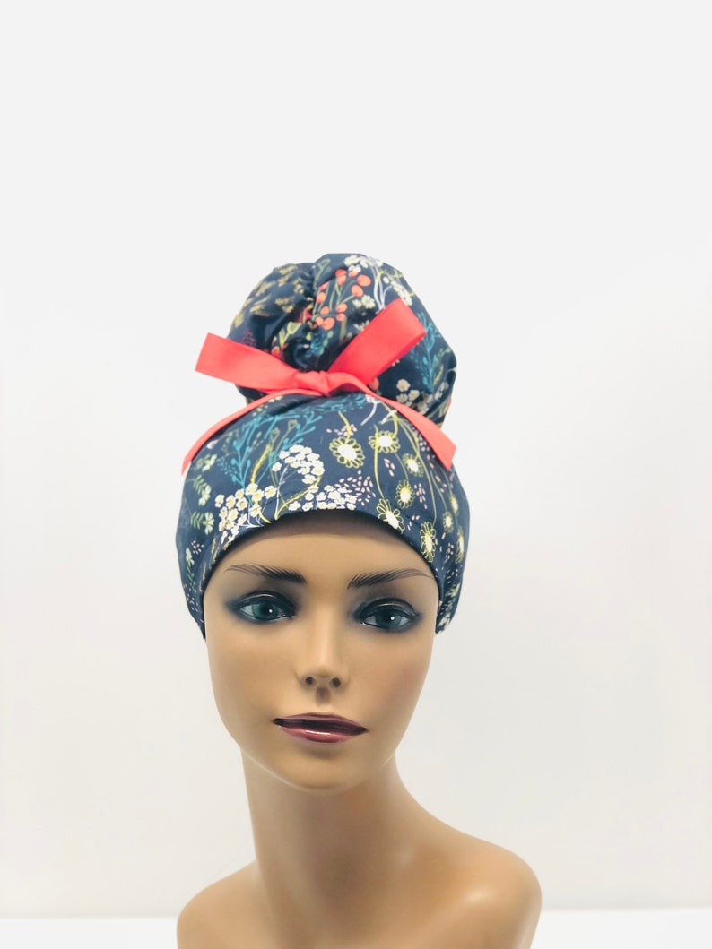 May include: A dark blue scrub hat with a floral print and a bright pink bow. The hat is designed to be worn with a bun.