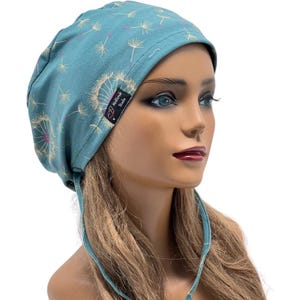 Peut inclure: Bonnet de gommage bleu sarcelle avec un motif de pissenlit. Le bonnet pr&eacute;sente un motif r&eacute;p&eacute;t&eacute; de t&ecirc;tes de graines de pissenlit en blanc et rose. Le bonnet porte une &eacute;tiquette &laquo;&nbsp;MediScrub Studio&nbsp;&raquo;.