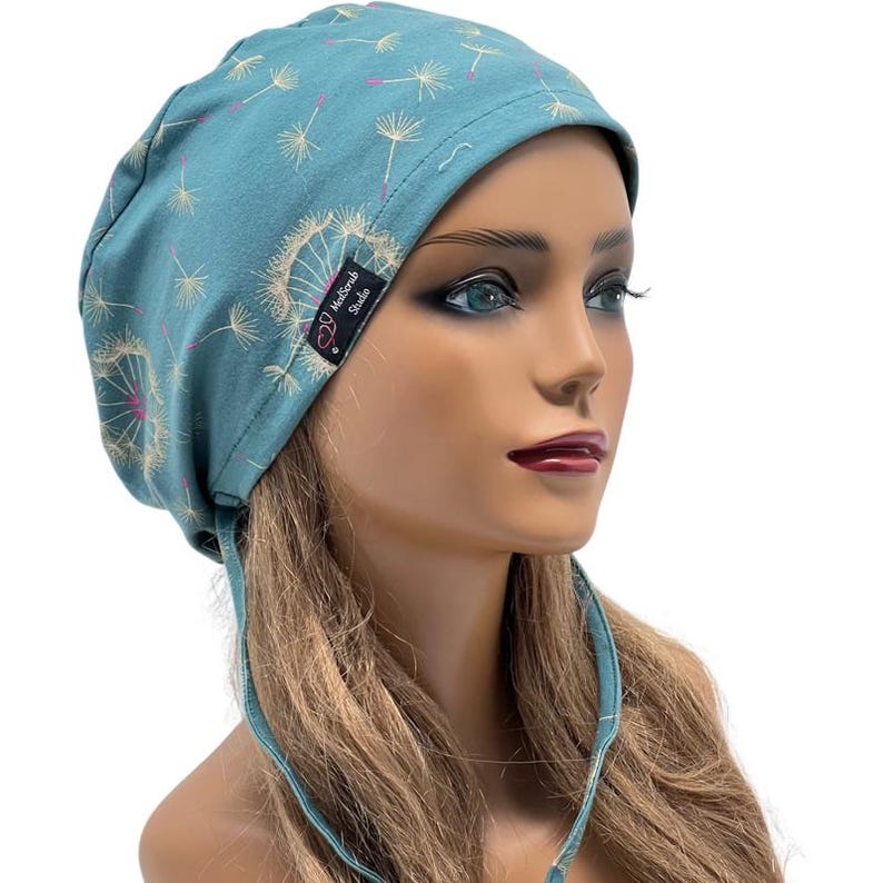 Peut inclure: Bonnet de gommage bleu sarcelle avec un motif de pissenlit. Le bonnet pr&eacute;sente un motif r&eacute;p&eacute;t&eacute; de t&ecirc;tes de graines de pissenlit en blanc et rose. Le bonnet porte une &eacute;tiquette &laquo;&nbsp;MediScrub Studio&nbsp;&raquo;.