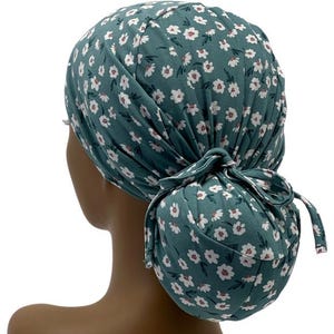 May include: Teal-colored scrub cap with a white floral pattern. The cap is tied in a bun at the back. The fabric is soft and gathered, with a bow detail. The cap is designed to cover the hair.