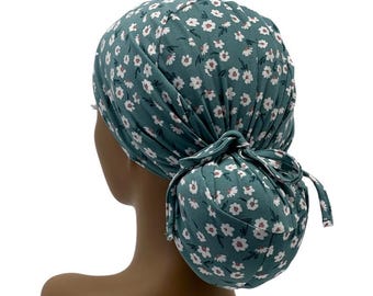 May include: Teal-colored scrub cap with a white floral pattern. The cap is tied in a bun at the back. The fabric is soft and gathered, with a bow detail. The cap is designed to cover the hair.