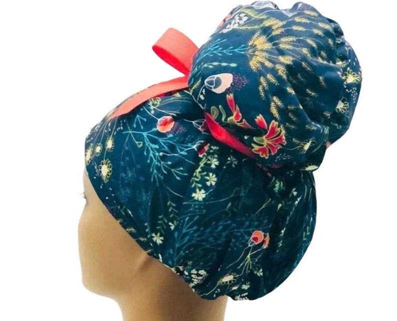 May include: A blue and green floral print surgical scrub hat with a red bow. The hat is designed to be worn with a bun.