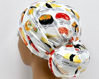 Könnte beinhalten: Eine weiße chirurgische Mütze mit einem bunten Sushi-Druck. Die Mütze zeigt verschiedene Arten von Sushi, darunter Uni, Toro, Tamago und Ebi. Die Mütze ist hinten zu einem Knoten gebunden.