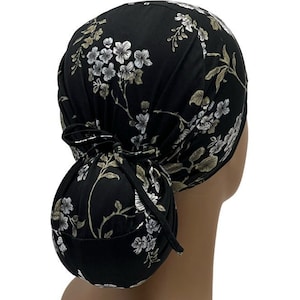 May include: Black scrub hat with a floral print featuring white and green flowers. The hat has a ponytail opening and ties at the back.