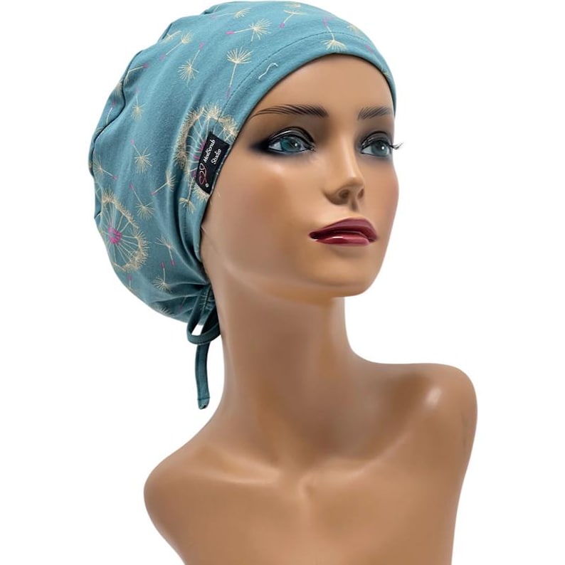 Peut inclure: Bonnet de gommage bleu sarcelle avec un motif de pissenlit. Le bonnet est dot&eacute; d'une attache dans le dos et d'une petite &eacute;tiquette noire avec du texte blanc. Le motif de pissenlit comprend des accents blancs et roses. Le bonnet est pr&eacute;sent&eacute; sur une t&ecirc;te de mannequin.