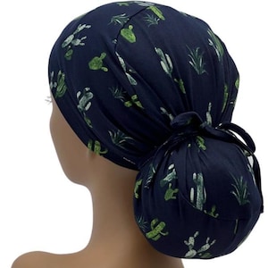 May include: A navy blue scrub cap featuring a pattern of green cacti and succulents. The cap is tied in a bun at the back. The fabric appears to be soft and comfortable, suitable for medical professionals or those in the service industry.