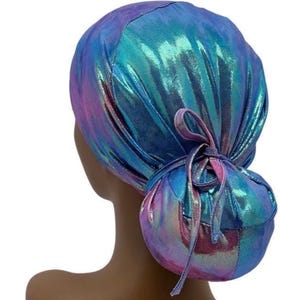 May include: A holographic, iridescent scrub cap in shades of blue, teal, and purple. The cap is gathered at the back and tied with a matching ribbon, creating a bun-like shape. The fabric has a shiny, metallic appearance.