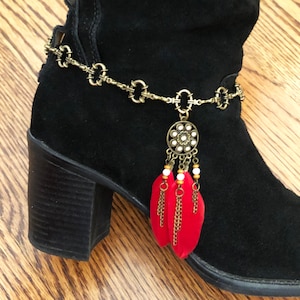 May include: A black suede boot with a decorative gold-colored chain ankle bracelet. The bracelet features a circular pendant with white accents and red feather charms. The boot has a block heel and is set against a wooden floor.