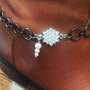Op de afbeelding: Een bruine leren laars met een zilveren ketting en een strass-bedel. De bedel heeft de vorm van een bloem met een parel die eraan hangt.