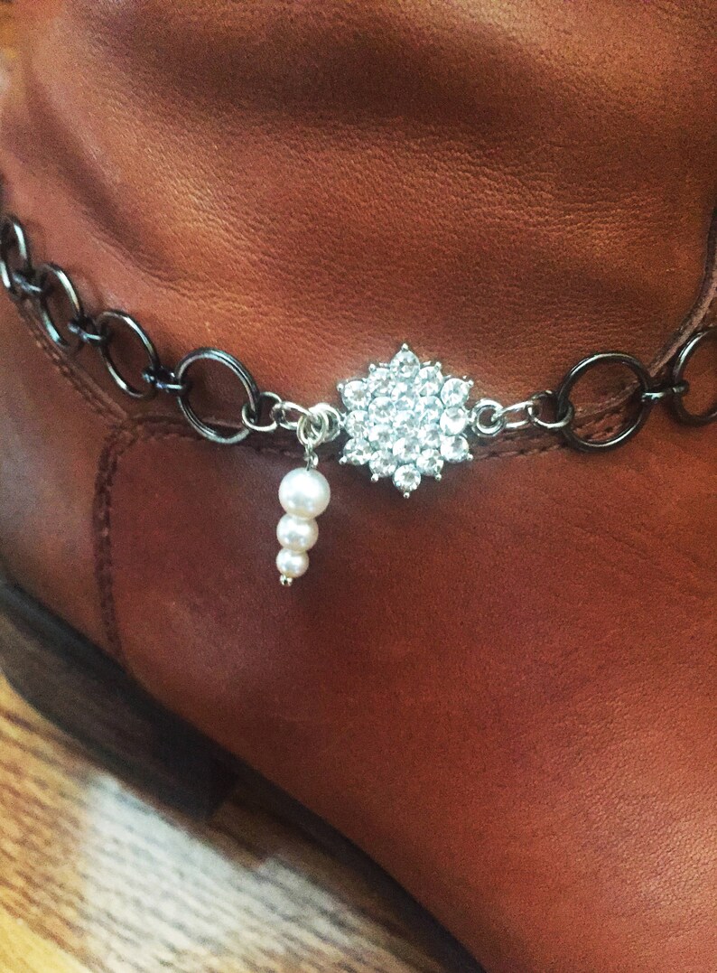 Op de afbeelding: Een bruine leren laars met een zilveren ketting en een strass-bedel. De bedel heeft de vorm van een bloem met een parel die eraan hangt.