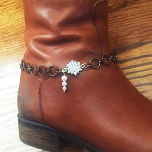 Op de afbeelding: Een bruine leren laars met een zilveren ketting en een strass-sneeuwvlok charme die eraan hangt. De charme heeft een parel-druppel.