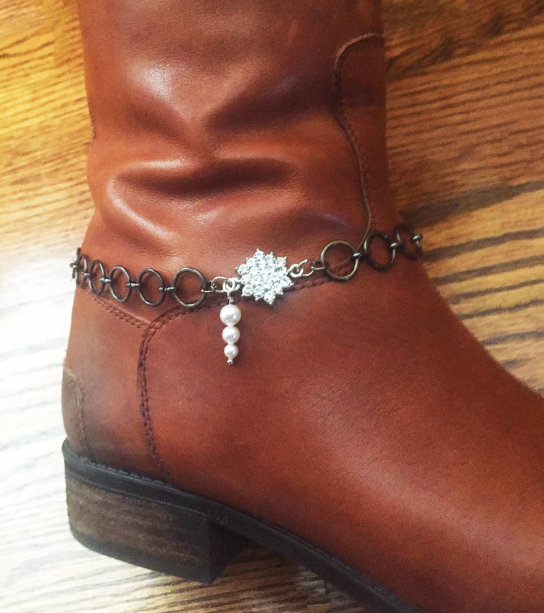 Op de afbeelding: Een bruine leren laars met een zilveren ketting en een strass-sneeuwvlok charme die eraan hangt. De charme heeft een parel-druppel.