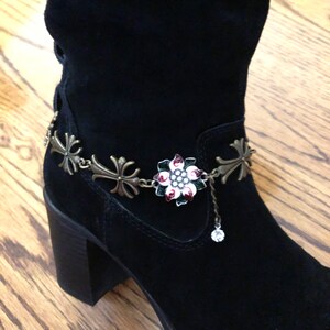 Puede incluir: Botas de gamuza negras con un brazalete de tobillo de cadena plateado y dorado con un dije de cruz y un dije de flor con esmalte rojo, blanco y verde.