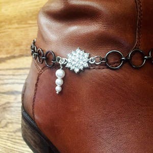Op de afbeelding: Een bruine leren laars met een zwarte ketting en een strass-bloemenbedel. Een streng parels hangt aan de bedel.